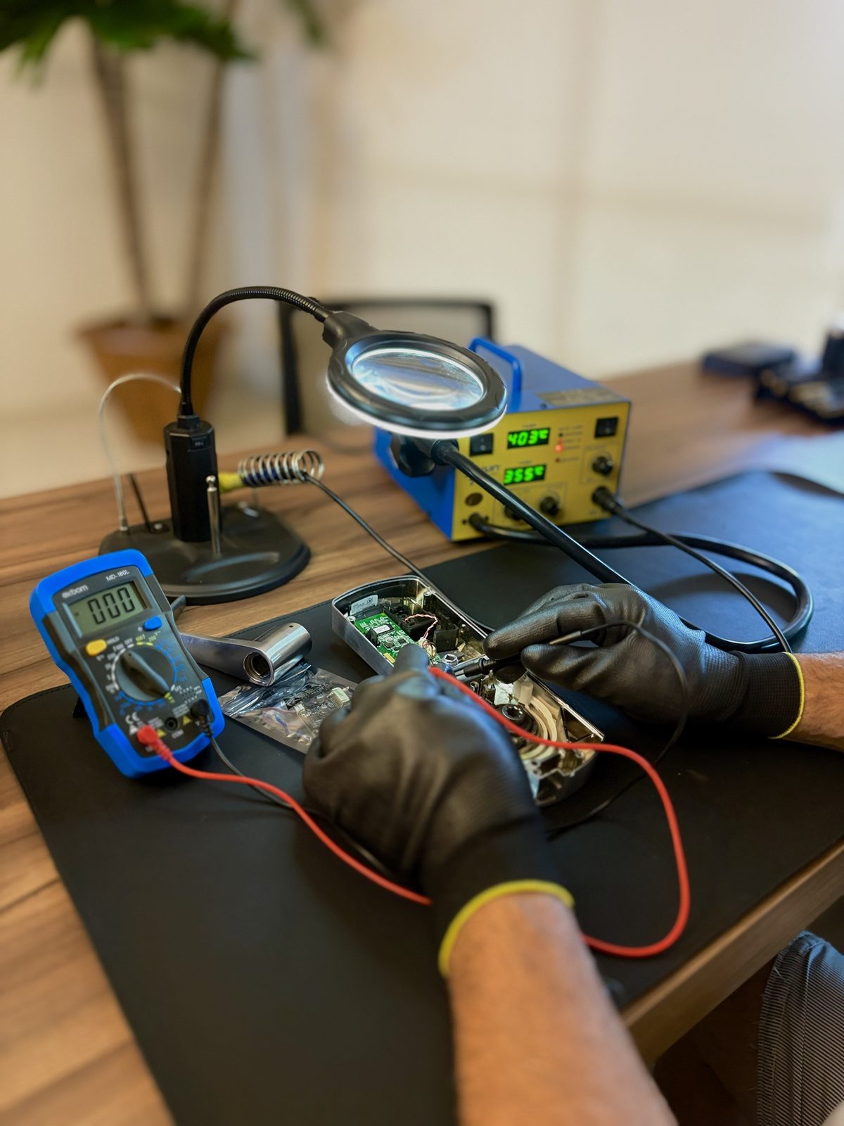 Técnico da RSV com luvas trabalhando na placa eletrônica de uma fechadura aberta na bancada, com multímetro, lupa iluminada, estação de solda e a placa RFID nova na embalagem antiestática.