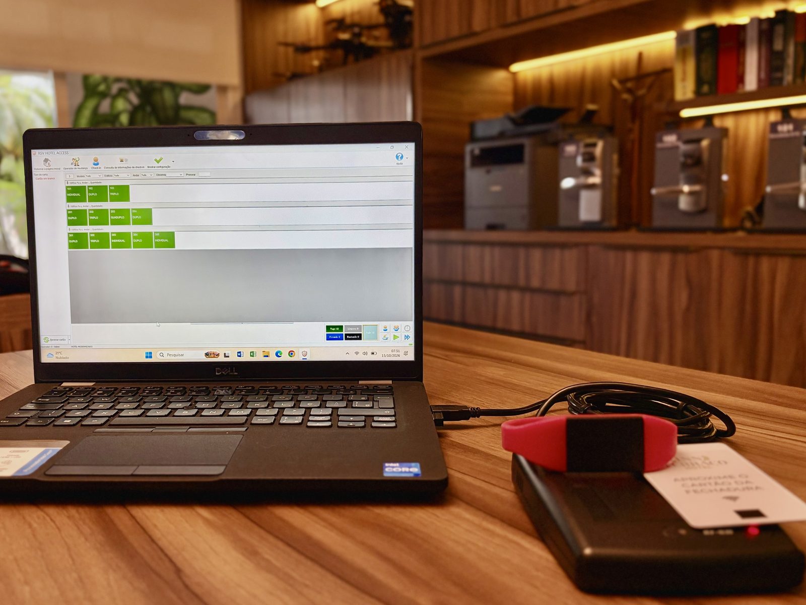 Notebook na recepção com o sistema RSV Hotel Access aberto no mapa de quartos, ao lado do encoder de mesa com um cartão e uma pulseira RFID.